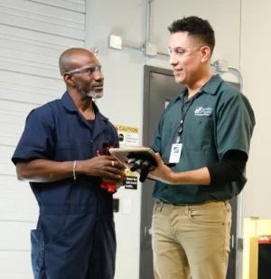 Two male PCSI employees in a warehouse review information on an iPad. The employee on the right holds the tablet while the employee on the left listens attentively.