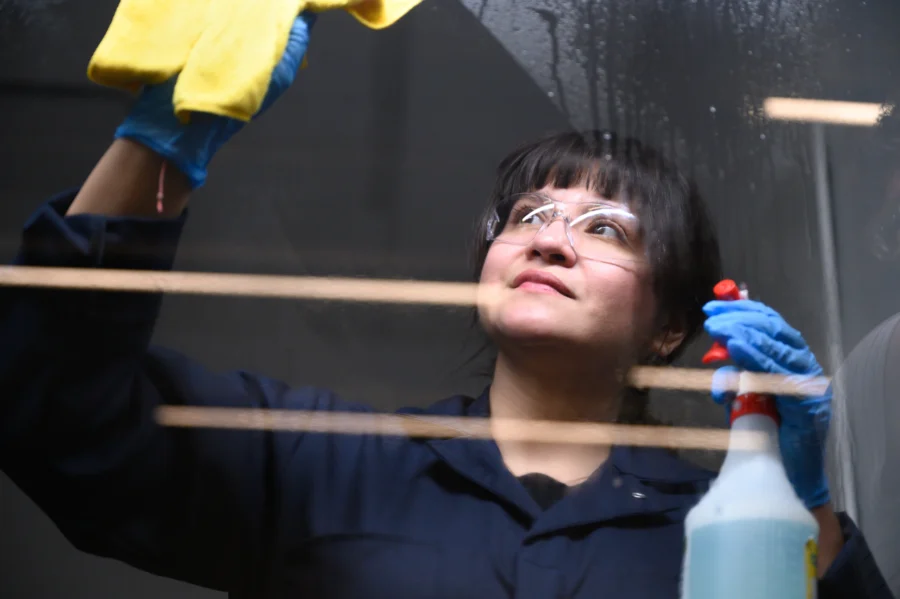 A young woman is shown cleaning a window. She is wearing overalls and protective eyewear.