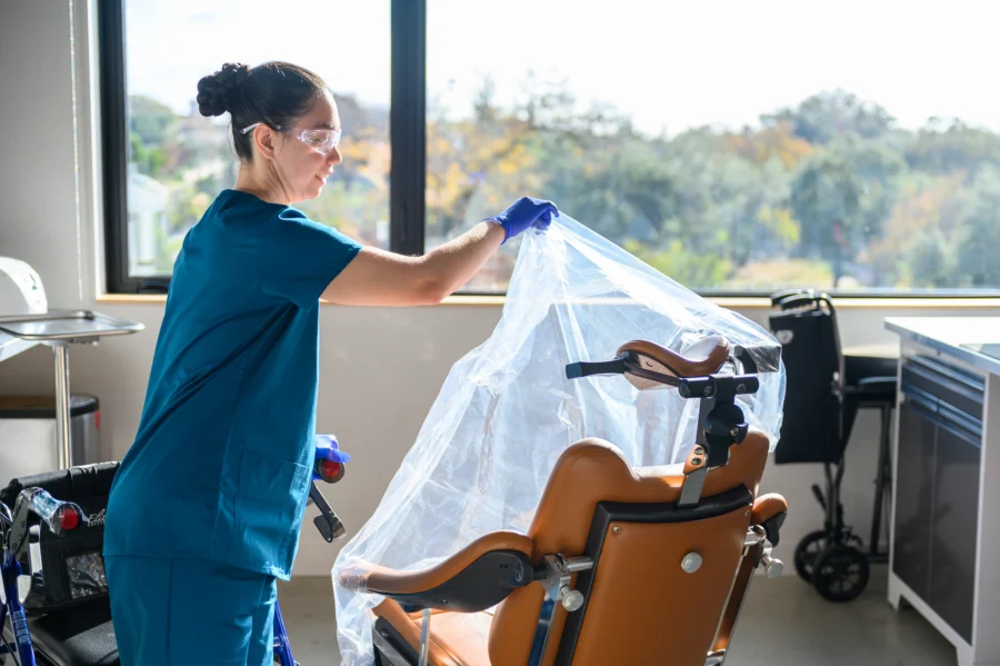 A female PCSI employee is shown covering medical equipment with a plastic sheet and wearing personal protective equipment while holding onto a rollator.
